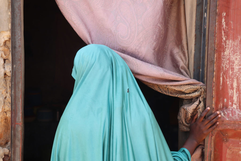 Person in turquoise garment, standing at doorway, holding curtain, partially hidden face.
