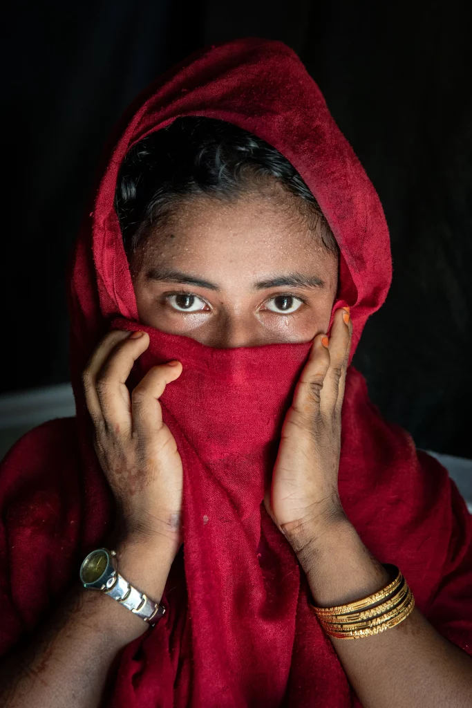 Woman with red scarf covering face, wearing bracelets and watch, looking directly at camera.