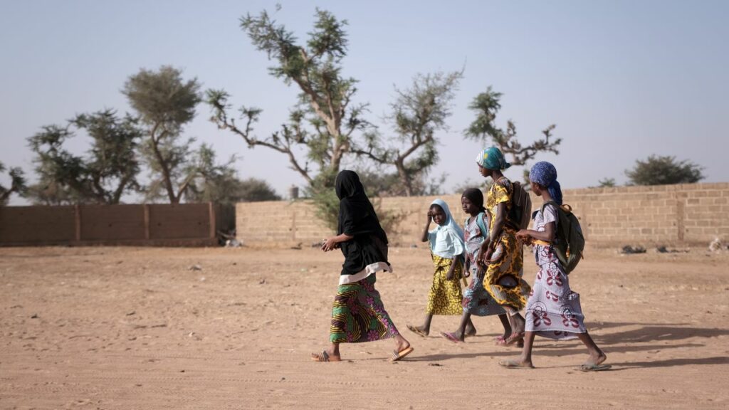 Children walk to school in a dry, rural area, wearing colorful clothing and carrying backpacks.