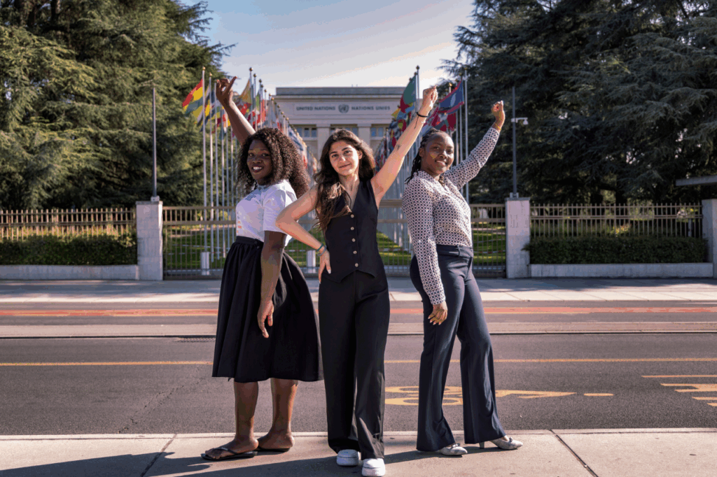 Three women confidently pose in front of the United Nations building with flag backdrop.