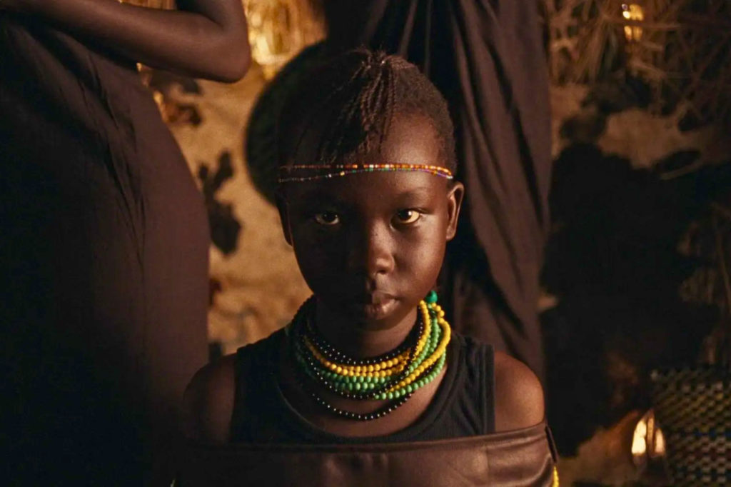 Child with colorful beads and braided hair in cultural attire, standing indoors with a thoughtful expression.