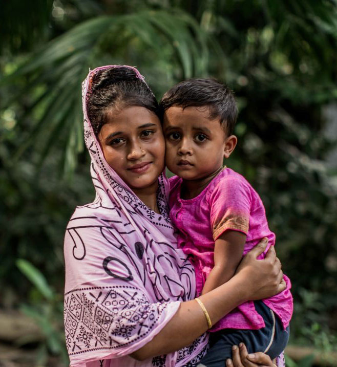 Mother holding child in vibrant pink clothing, surrounded by lush greenery, smiling at the camera.
