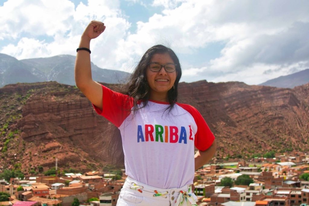 Smiling woman in an ARRIBA shirt raises fist triumphantly against a scenic mountain backdrop.