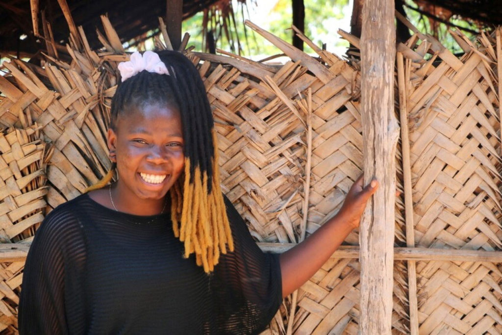 Smiling woman with dreadlocks standing by woven wooden wall in sunlight.