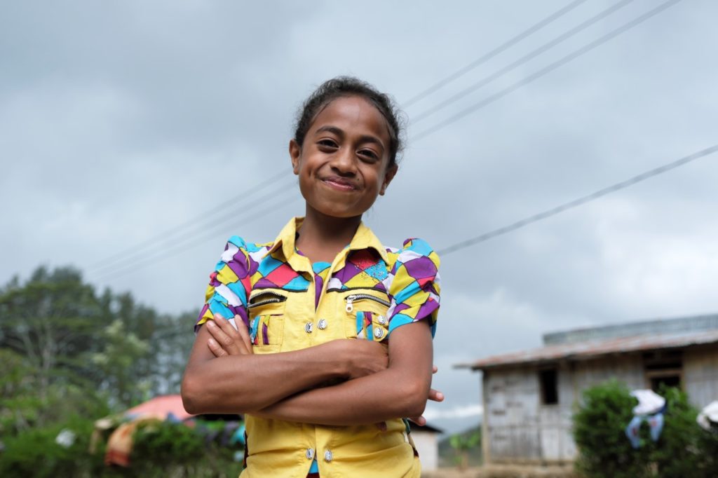 Smiling girl in colorful shirt stands confidently outdoors, cloudy sky and rustic house in the background.