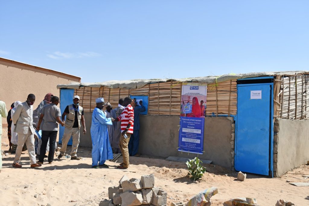 Community members gather outside a local building for an aid event, with a blue door and informational posters.