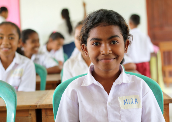 Smiling student in classroom wearing a name tag, sitting in front of classmates.