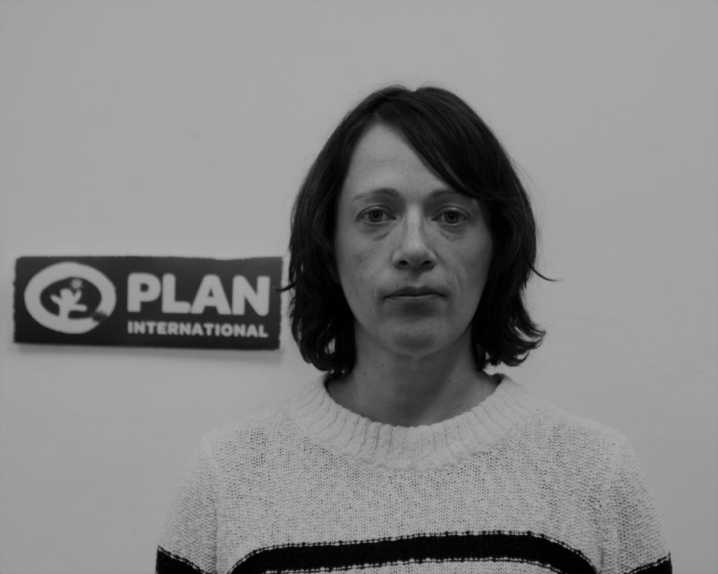 Individual in a white sweater stands before a Plan International sign, black and white photo.
