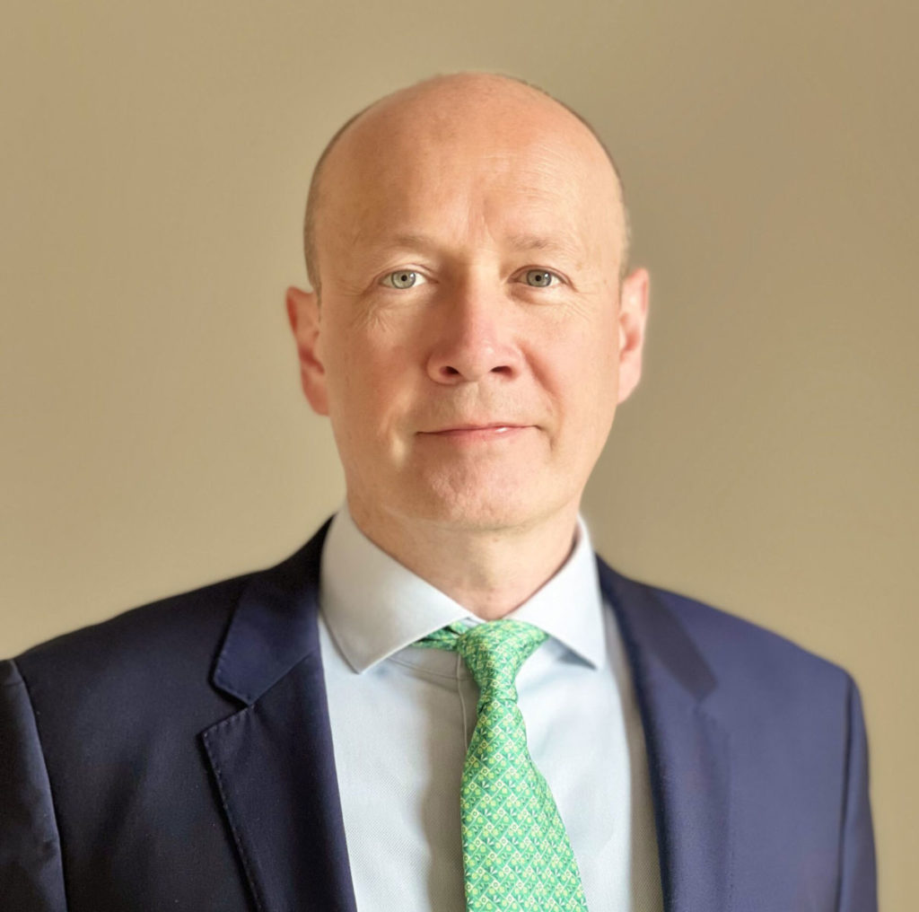 Bald man in a suit with a light green tie, standing against a neutral background. Formal attire.