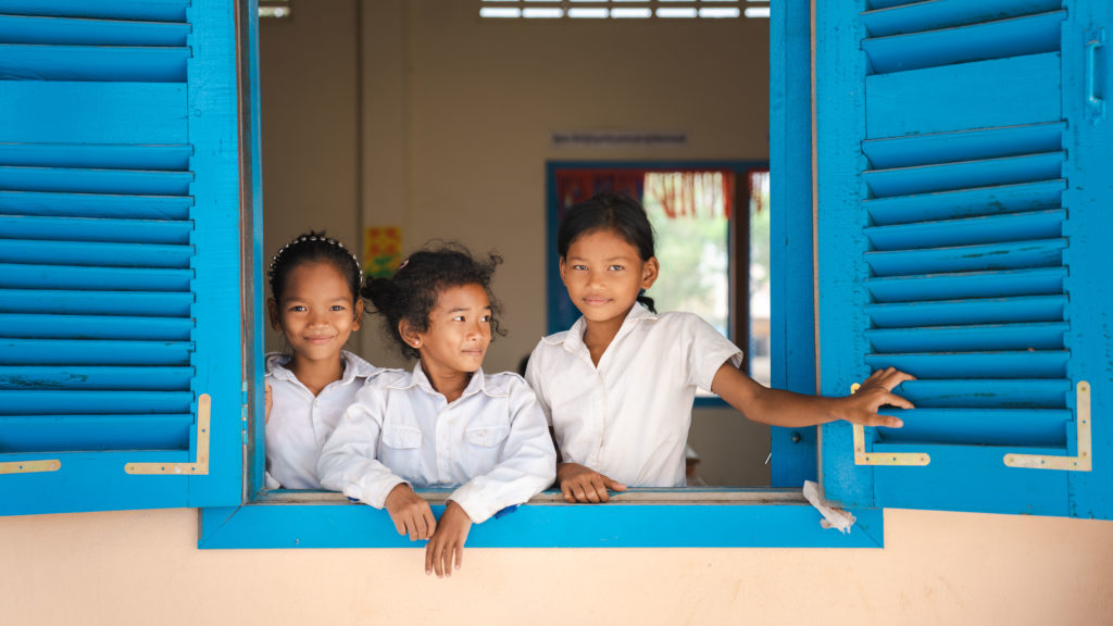 Three children in Cambodia dressed in school uniforms look outside their school window