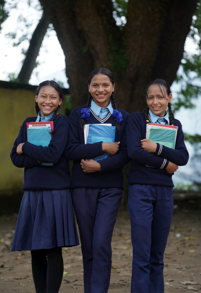 Three girls wearing school uniforms hold books and smile at the camera