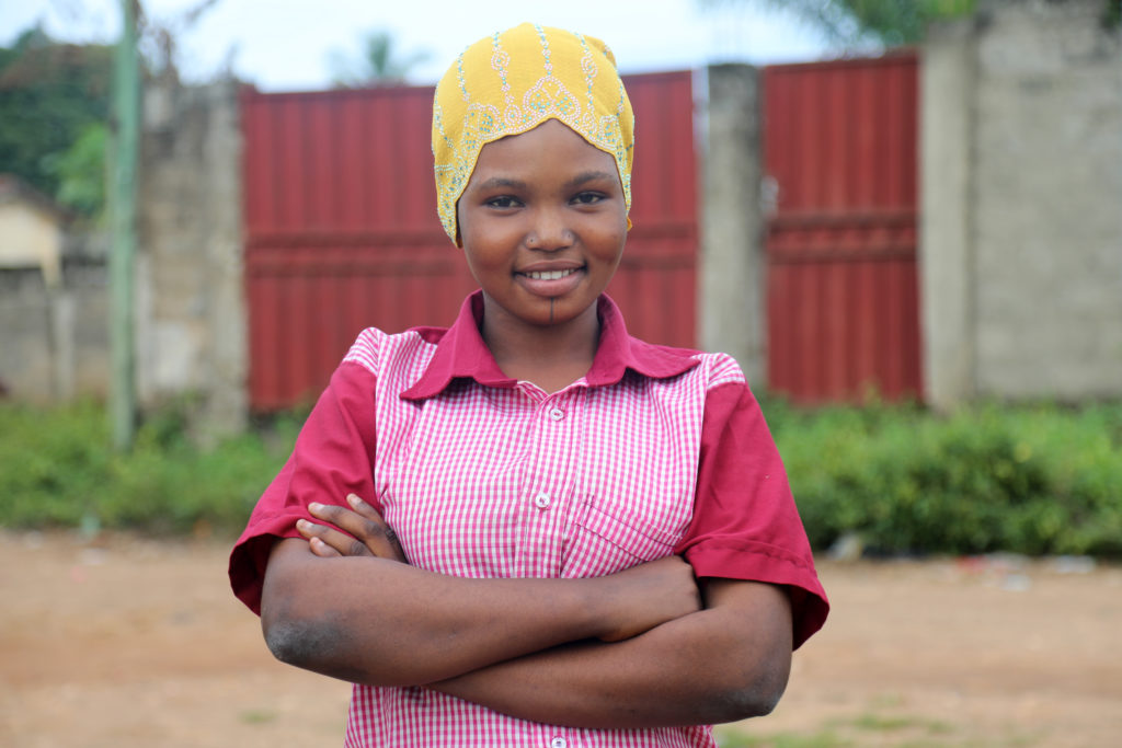 A girl in a pink top and yellow handkerchief looks at the camera with her arms crossed