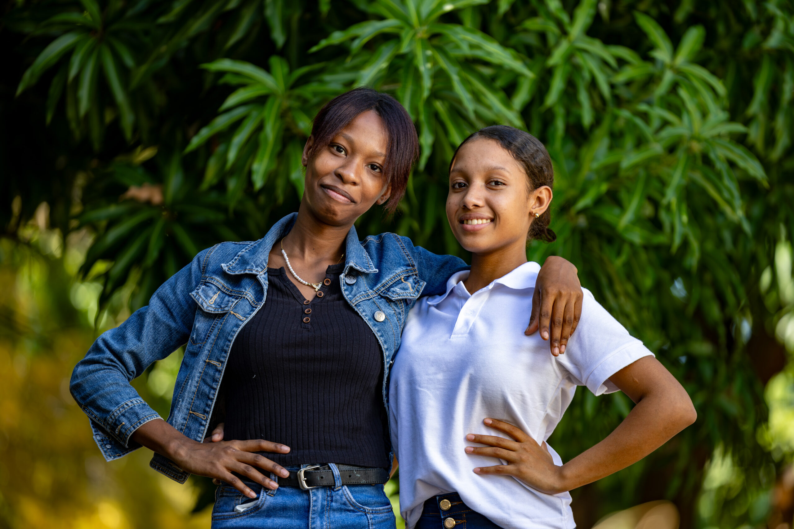 Two girls from the Dominican Republic, one dressed in a denim jacket and one in a white shirt, stand together smiling.