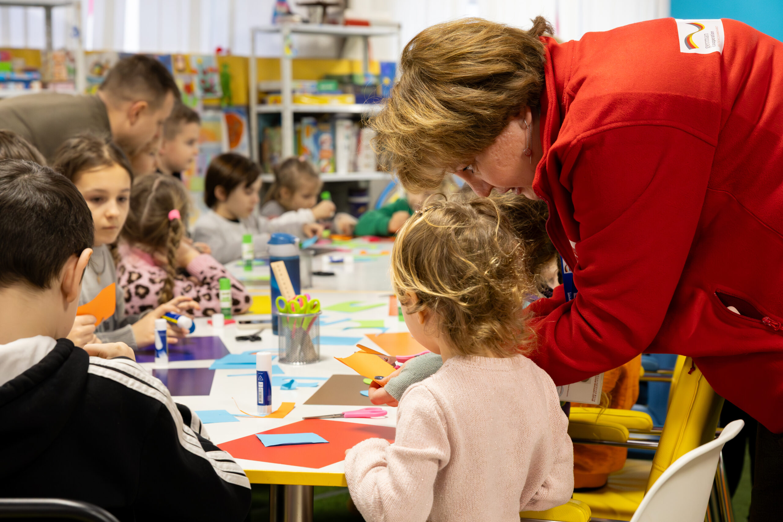A woman in a red jacket helps a child with an art project