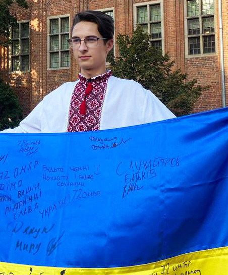 A young man dressed in a traditional Ukrainian shirt, holds a Ukrainian flag