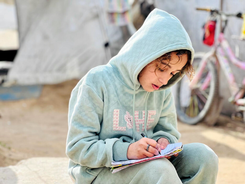 A girl in a light green sweatshirt with hood writes in a journal