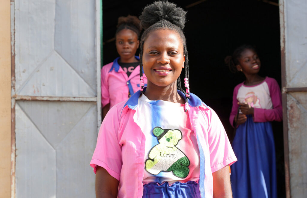 A girl in a pink shirt with a blue collar stands in front of the camera smiling