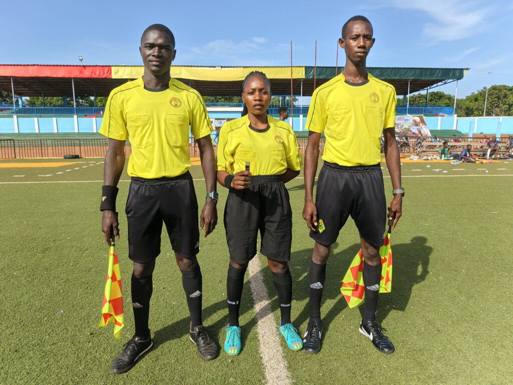 Three referees stand in a field, the middle one is a woman, and the other two are men